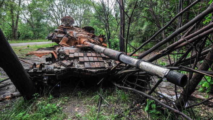 A destroyed tank rests on fallen trees ten yards or so off the shoulder of a tree-lined road.