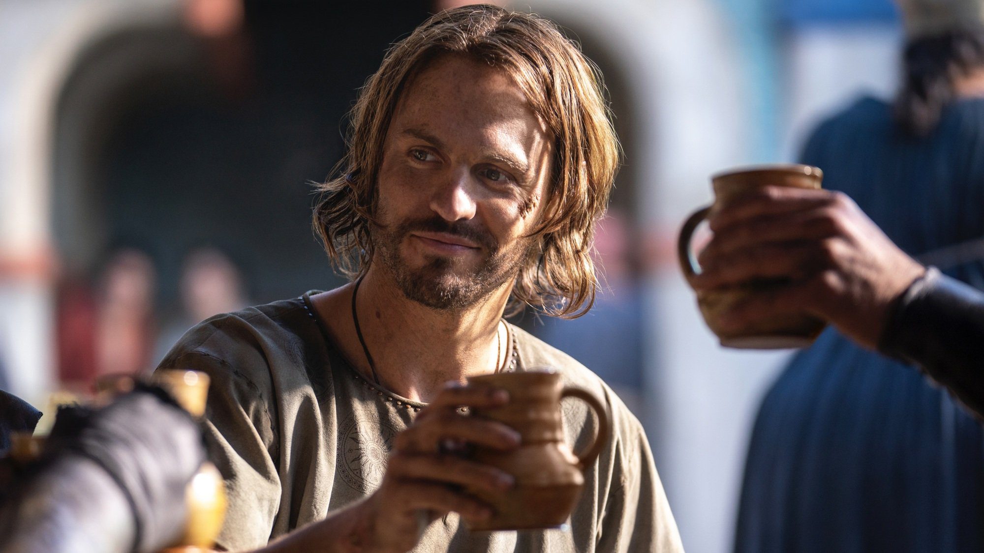 A man with brown hair of medium length raises a brown ceramic mug in a toast.